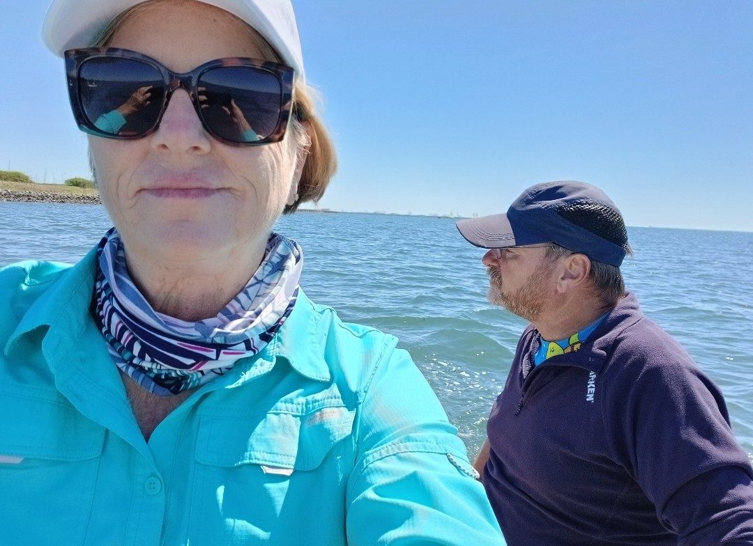Charlotte out on the water in a white cap and sunglasses, a patterned neck sock worn around her neck, with Dave behind her